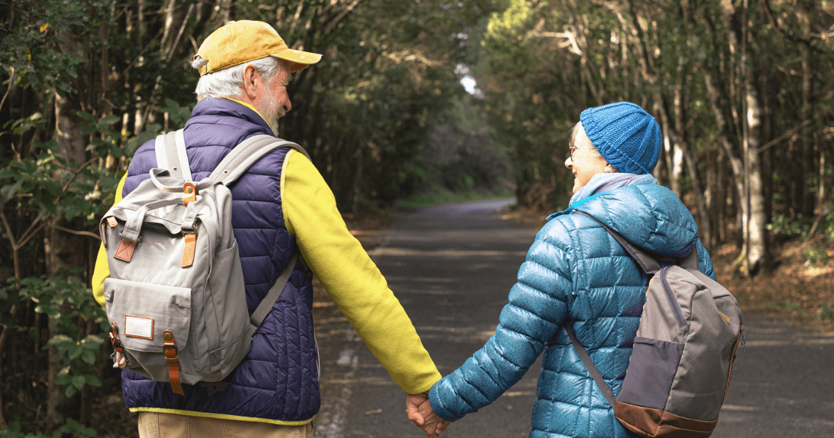 Senior couple with backpacks on a walk outdoors surrounded by trees in a path during Retirement in Hillsborough Township