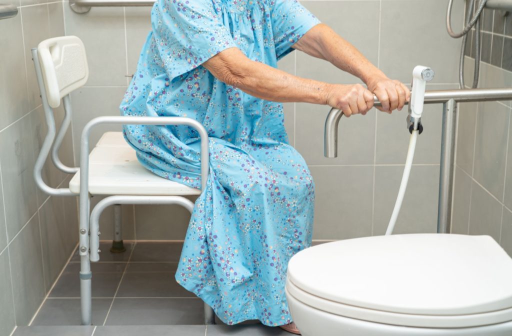 Elderly person seated on a bath bench holding a safety rail in an accessible bathroom for daily living support.