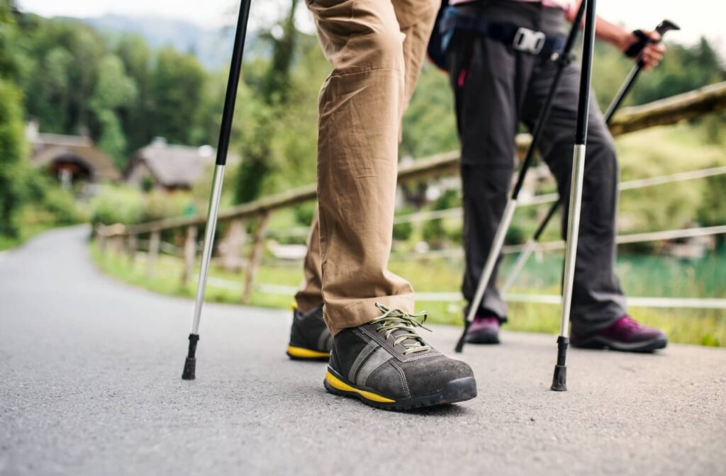 Close-up of two people walking with trekking poles on a paved path showing supportive walking shoes in use outdoors.