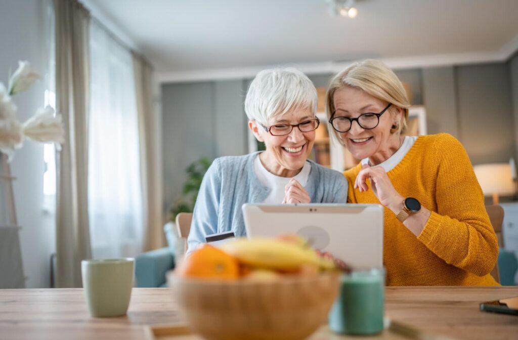 Two people smiling and looking at a tablet during a video call while sitting at a table with fruit and coffee cups nearby.