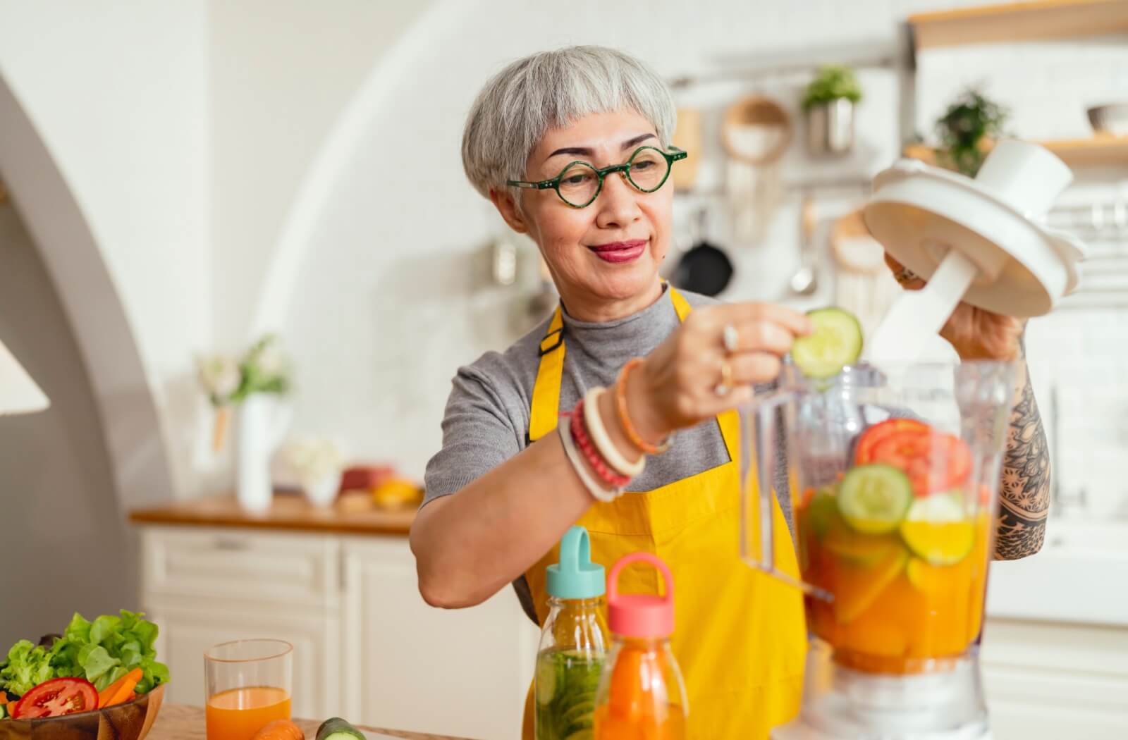 In a brightly lit kitchen, a senior places fruits and vegetables into a blender to make a healthy juice to drink