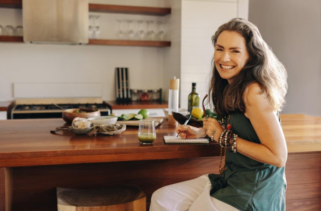 A happy senior sits at a kitchen table with a pen and paper to make plans for meals for the week, to ensure nourishing meals