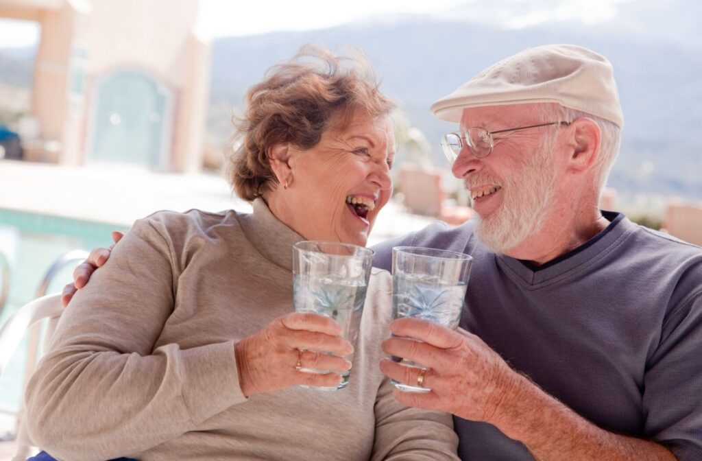 A senior couple laughing, each holding a tall glass of water to stay hydrated while spending some time outdoors