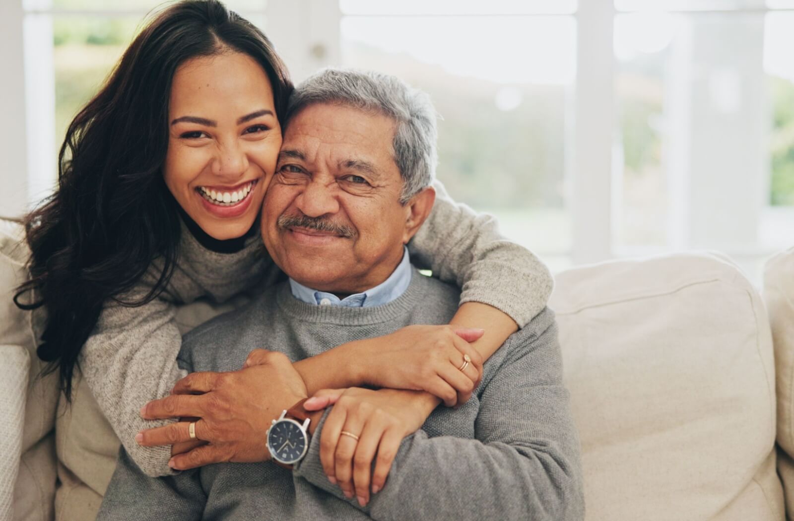 An adult hugs their seated older parent over the shoulders, both smiling from the joy and love their connection brings