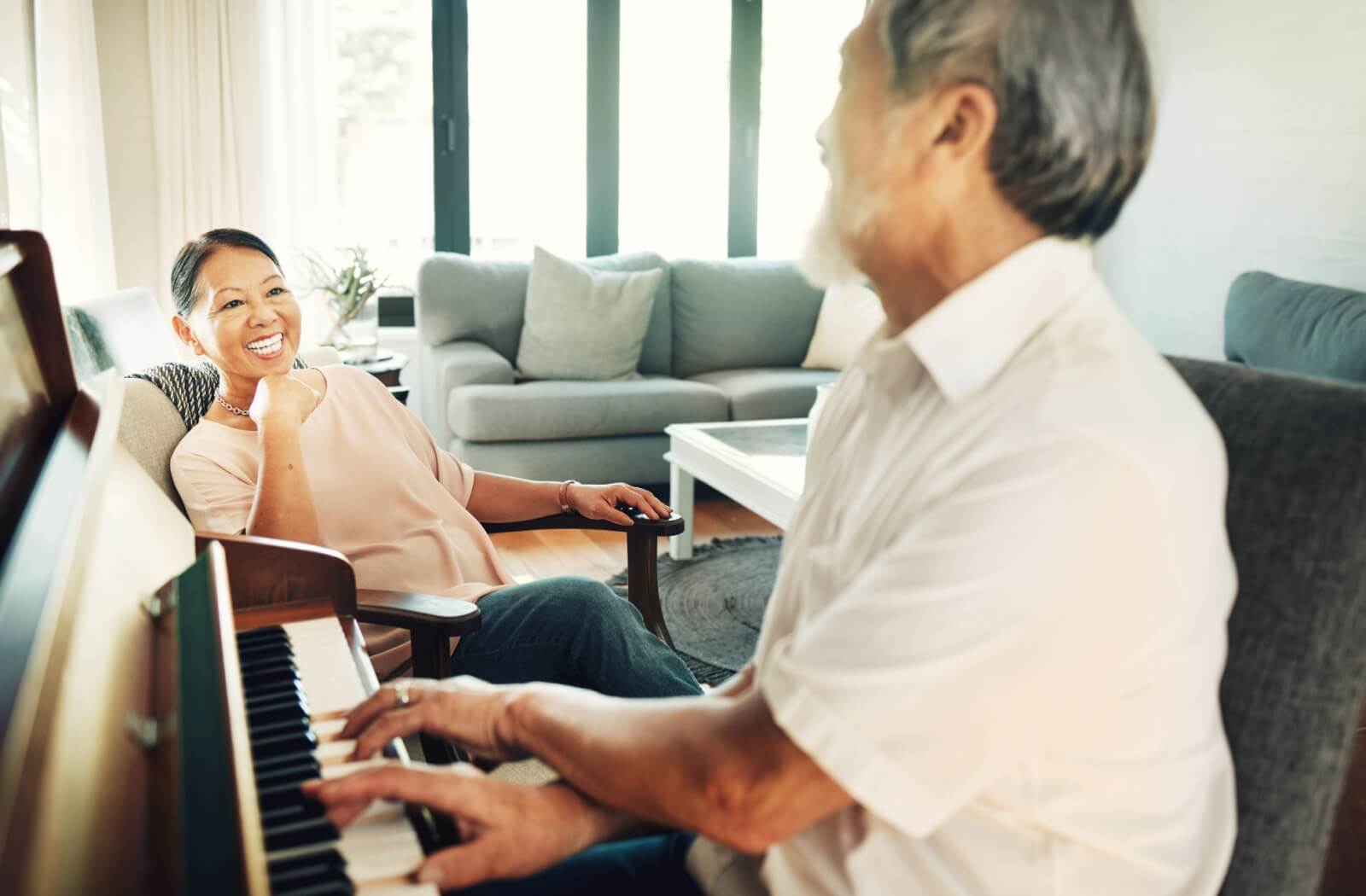 A smiling senior sits and listens to their partner with dementia play the piano as part of their daily routine
