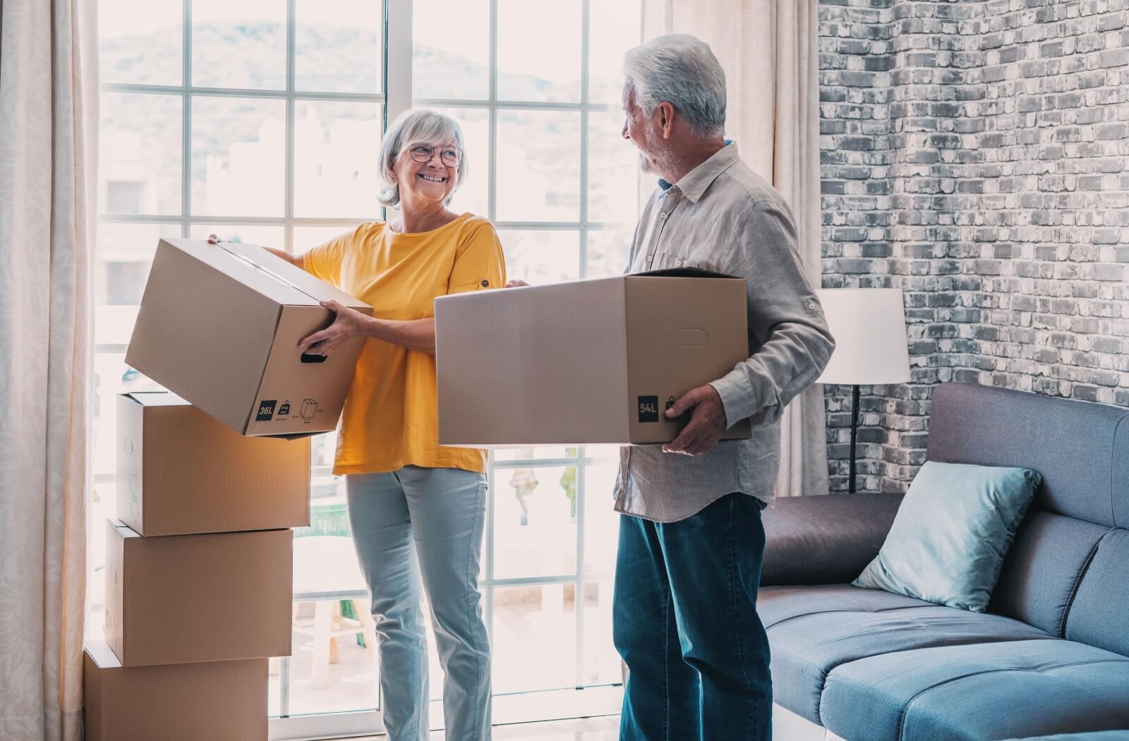 A senior couple holding moving boxes smiles at each other as they settle into their new senior living community suite