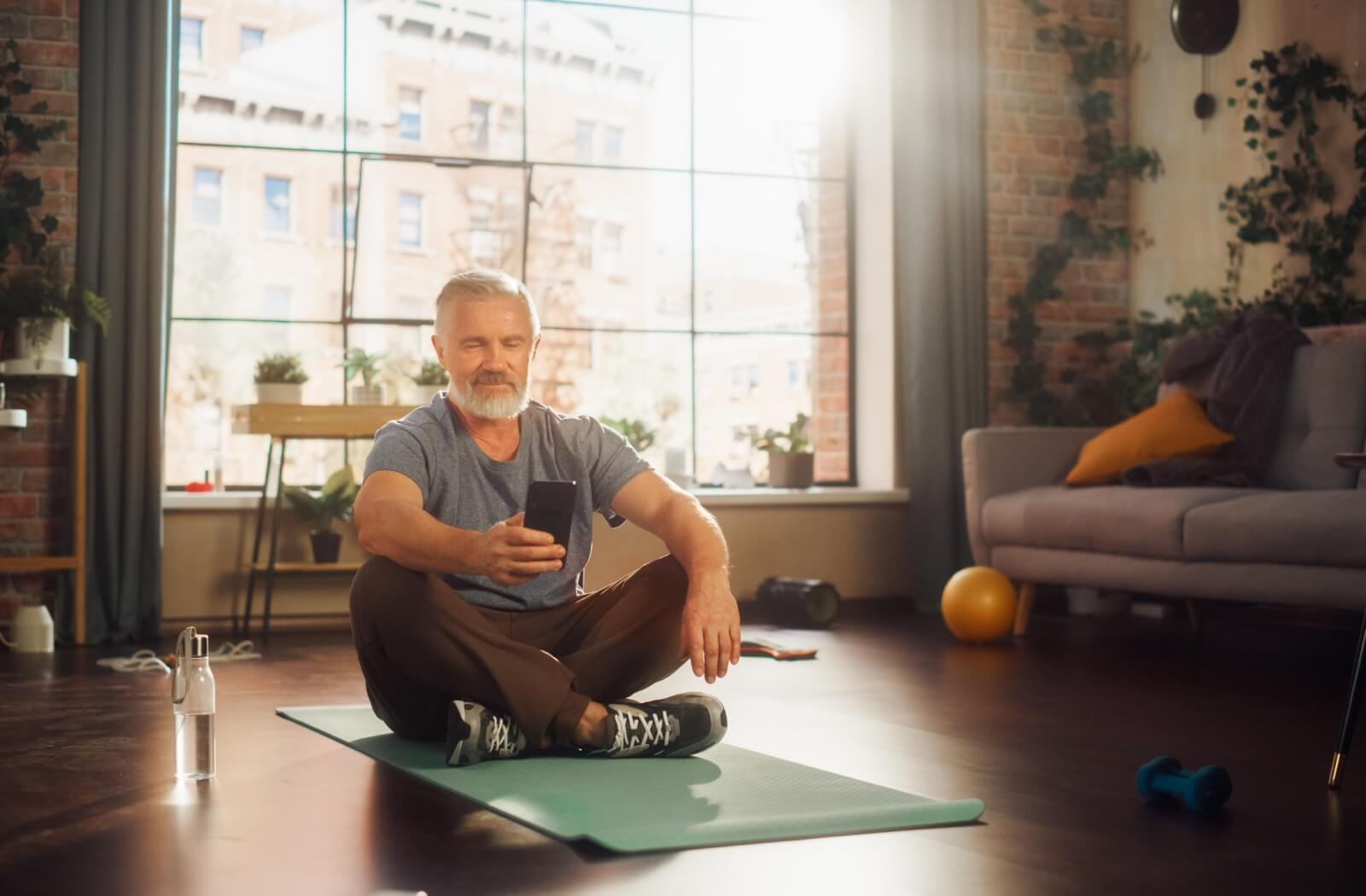 A senior sitting on a yoga mat on the floor uses their phone to pull open a yoga app to help with their physical health