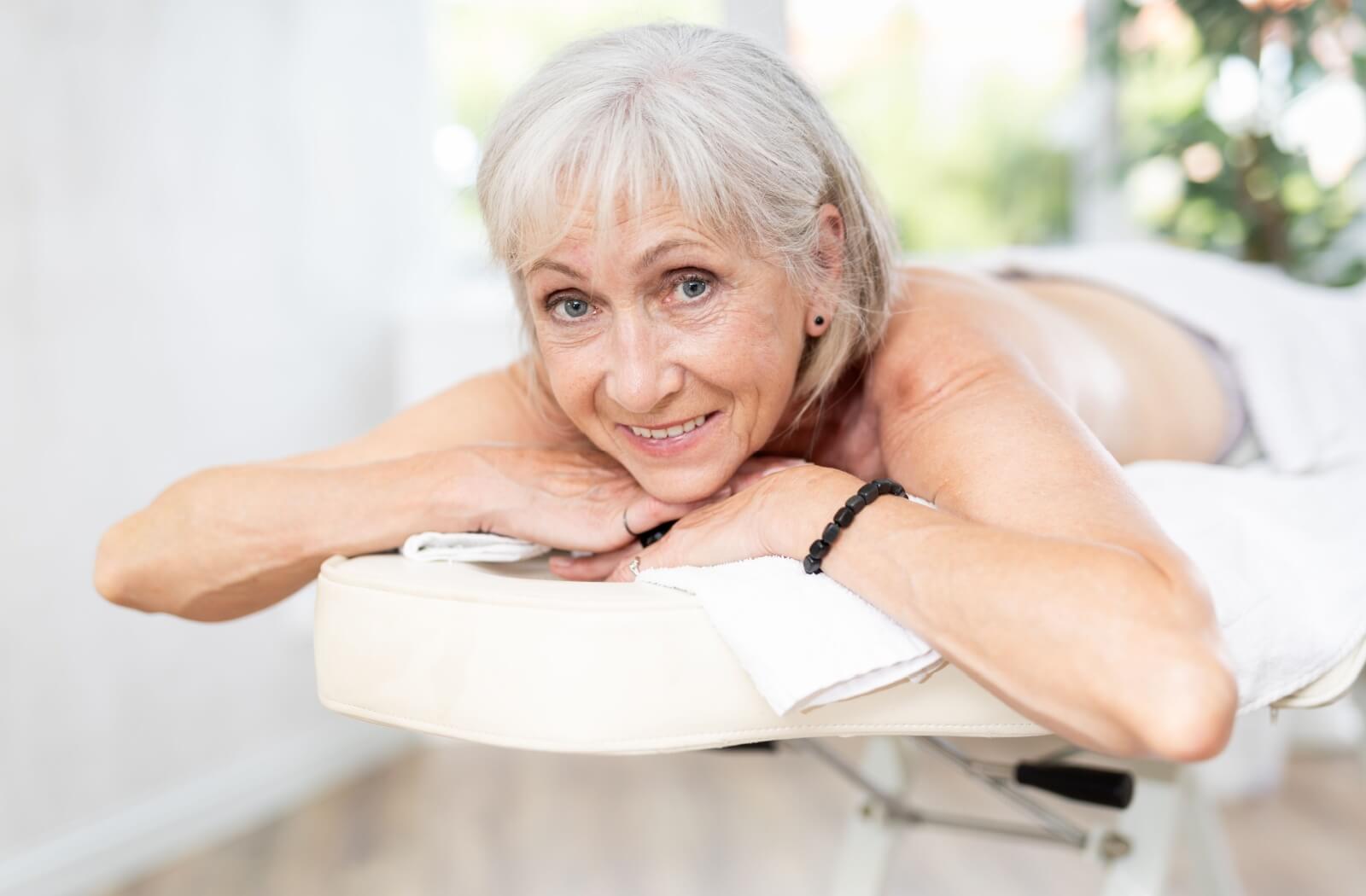 A grey haired person is lying on their stomach on a massage table, with their hands propping up their chin. They are smiling at the viewer and are relaxed.