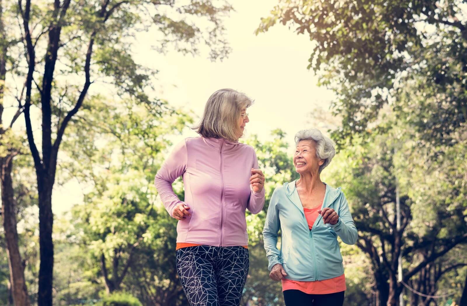 Two people walking and smiling in a sunny park wearing activewear surrounded by trees and greenery