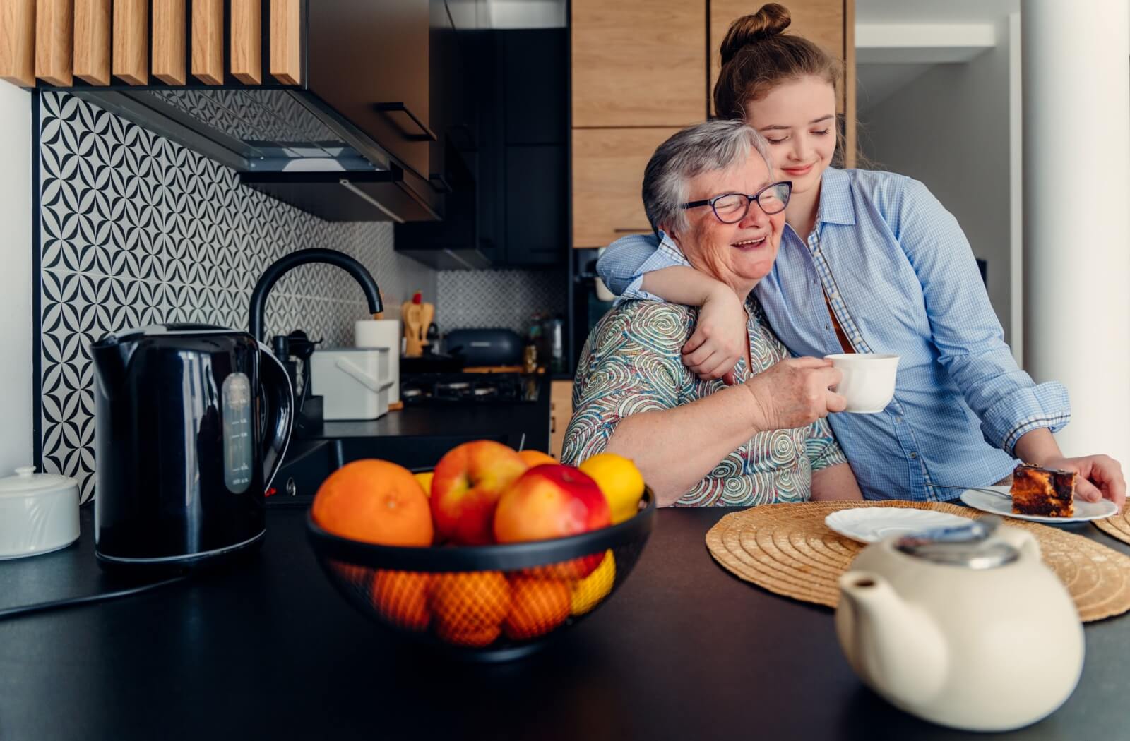 Two people share a hug in a kitchen while enjoying tea and dessert with a bowl of fruit and teapot on the counter