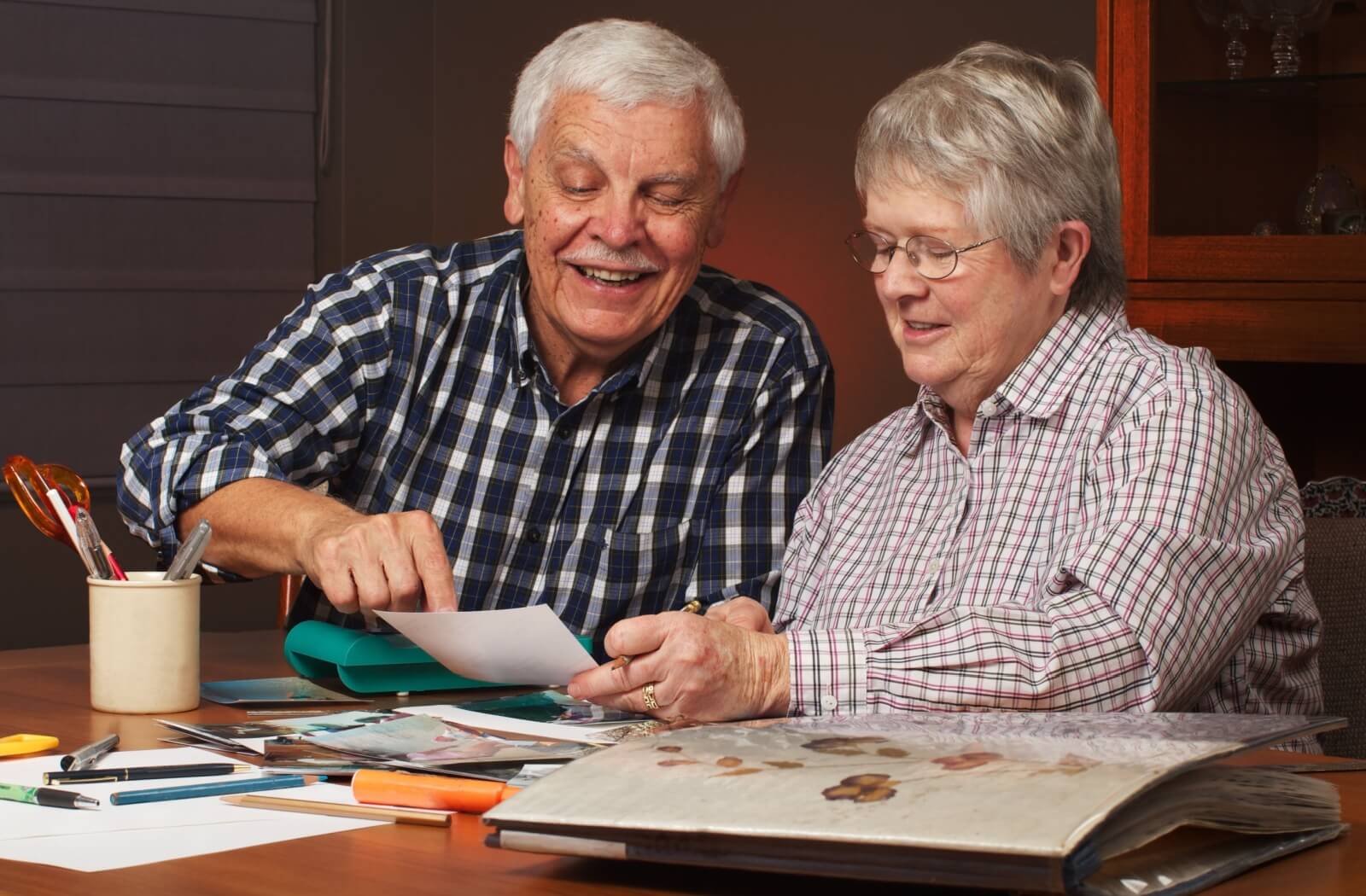 Senior couple sitting at a table scrapbooking with photos and colored pens while smiling and engaged in conversation