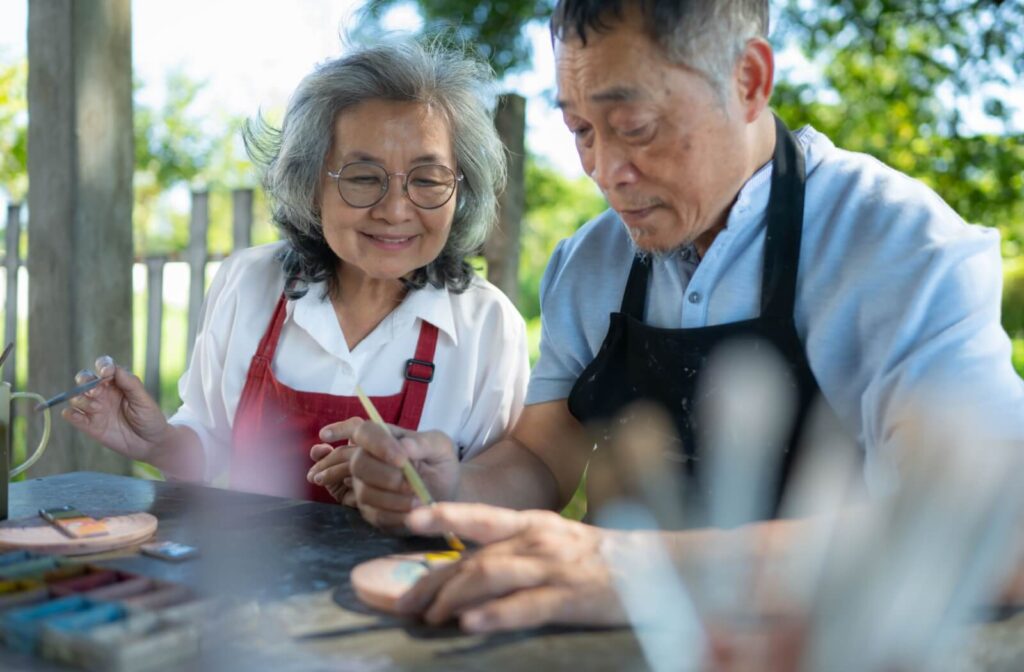 Older adults painting together outdoors in aprons while smiling and focused in a sunlit garden setting